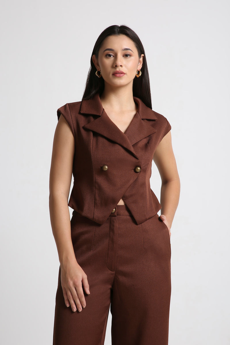 Female model wearing a brown sleeveless double breasted blazer top with wide lapels and matching high waist trousers from Muzera, posing in a studio close up front shot.